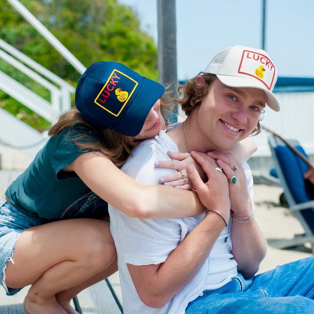 Couple wearing navy and white Lucky Duck trucker hats at sunny beach lifestyle shot