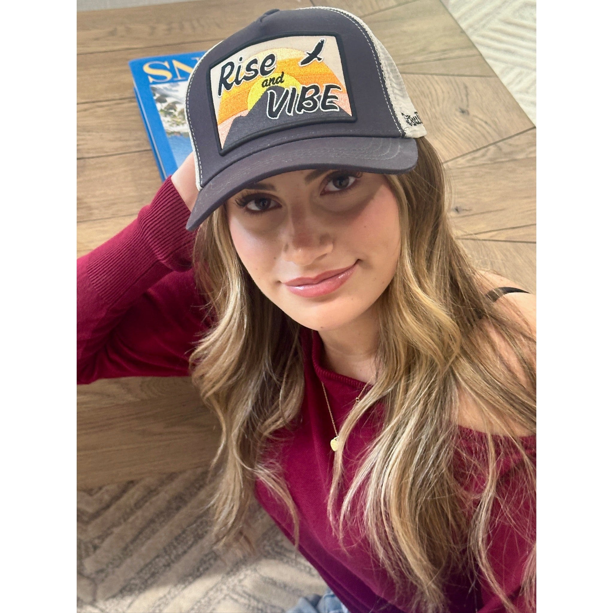 Woman wearing charcoal Rise & Vibe trucker hat with sunset mountain patch and eagle design